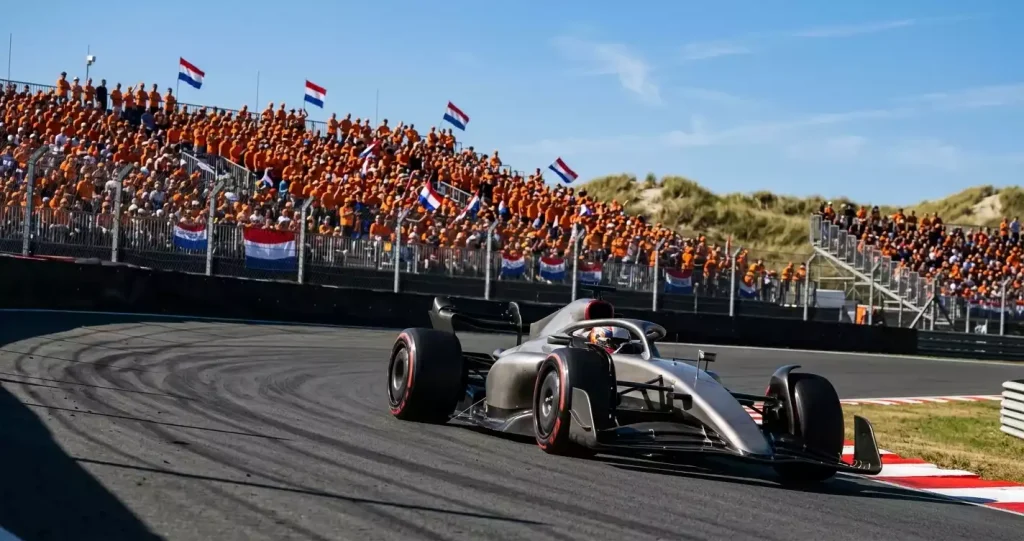 Formule&nbsp;1-wagen in de hooggebankte Hugenholtzbocht op het circuit van Zandvoort met oranje publiek op de tribunes en duinen op de achtergrond